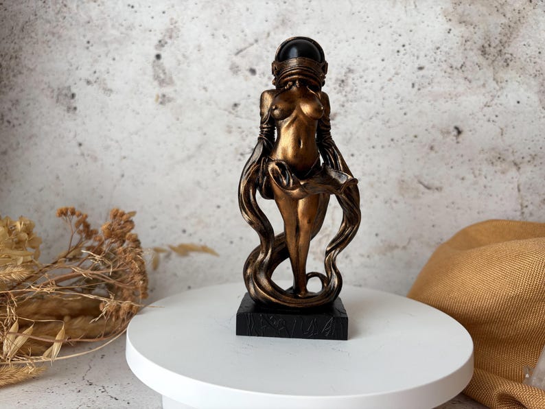 May include: A bronze-colored sculpture of a female figure with a helmet-like head covering and flowing drapery. The statue is mounted on a black rectangular base and stands on a white surface. The figure's form is stylized and elegant.