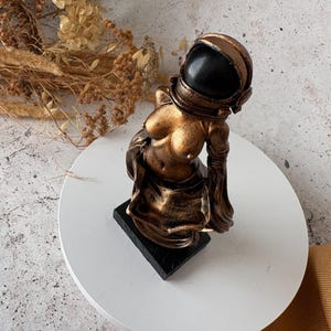 May include: A bronze-colored astronaut sculpture with a black helmet. The figure is posed with a contemplative expression, mounted on a black rectangular base. The sculpture has a metallic finish and is set against a neutral background.