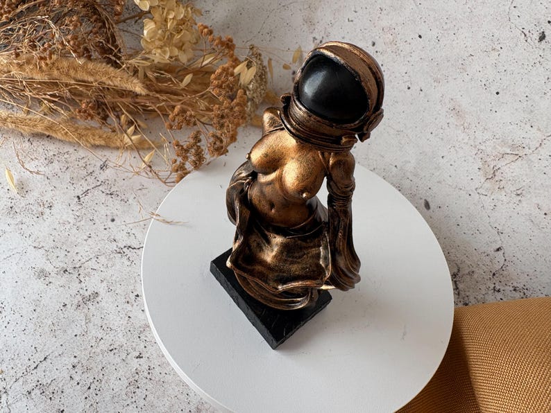May include: A bronze-colored astronaut sculpture with a black helmet. The figure is posed with a contemplative expression, mounted on a black rectangular base. The sculpture has a metallic finish and is set against a neutral background.