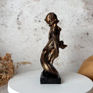 May include: A bronze-colored sculpture of a figure wearing a diving helmet and flowing fabric. The statue is mounted on a black rectangular base. The figure's form is stylized, with a focus on the helmet and the draping of the fabric.