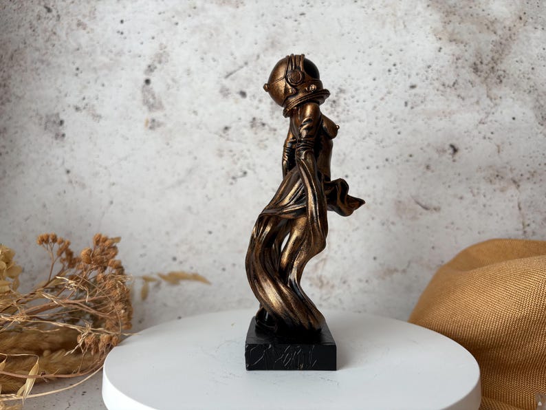 May include: A bronze-colored sculpture of a figure wearing a diving helmet and flowing fabric. The statue is mounted on a black rectangular base. The figure's form is stylized, with a focus on the helmet and the draping of the fabric.