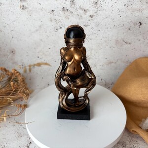 May include: A bronze-colored statue of a figure wearing an astronaut helmet. The statue is on a black base and stands on a white circular surface. The figure's body is partially visible, with flowing drapery. The background is a mottled gray and white.