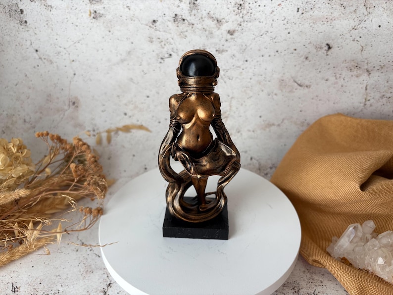 May include: A bronze-colored statue of a figure wearing an astronaut helmet. The statue is on a black base and stands on a white circular surface. The figure's body is partially visible, with flowing drapery. The background is a mottled gray and white.