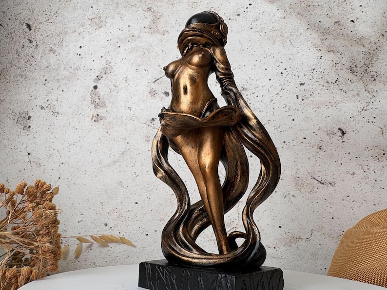 May include: A bronze-colored statue of a woman with long flowing hair and a helmet. The statue has a black base and is set against a neutral background. The figure's form is stylized, with a focus on flowing lines and curves.