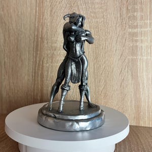 May include: A silver-colored figurine of a woman and a horned figure embracing. The figurine is standing on a round, silver-colored base.