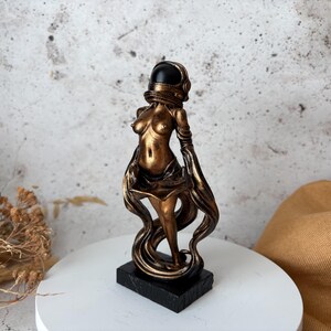 May include: A bronze-colored sculpture of a woman wearing an astronaut helmet. The figure is standing on a black rectangular base. Flowing fabric-like elements cascade down from the figure's arms and body. The sculpture is displayed on a white circular surface.