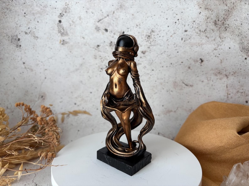 May include: A bronze-colored sculpture of a woman wearing an astronaut helmet. The figure is standing on a black rectangular base. Flowing fabric-like elements cascade down from the figure's arms and body. The sculpture is displayed on a white circular surface.