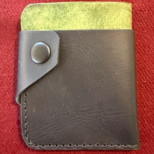 Puede incluir: Una cartera de cuero negro con interior verde. La cartera tiene un cierre de botón a presión y está cosida alrededor de los bordes. La cartera es rectangular y parece estar hecha de materiales de alta calidad.