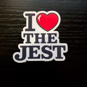 May include: A white sticker with the text "I THE JEST" in a bold, black font. A red heart with a white highlight replaces the word "LOVE". The sticker is on a black surface.
