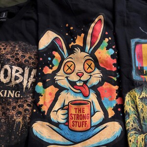 May include: A black t-shirt featuring a graphic of a cartoon rabbit with X-shaped eyes and a tongue sticking out, sitting cross-legged while holding an orange mug that reads THE STRONG STUFF, set against a colorful, splattered paint background.