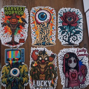 May include: A collection of six colorful stickers with unique, spooky designs. The stickers feature images of a scarecrow, an eyeball, a rose, a television head, a clown, and a girl holding a heart. The stickers have the text "Harvest Horror" and "Lucky Sammy".