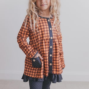 Könnte beinhalten: Eine langärmelige Tunika mit orange-schwarzem Karomuster, einer dunkelblauen Knopfleiste mit weißen Punkten sowie einer passenden dunkelblauen Tasche und einem gerüschten Saum mit Punkten. Das Kleidungsstück ist mit hellen Knöpfen versehen.