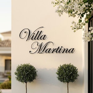May include: Exterior shot of a building with a white wall featuring the black cursive text "Villa Martina." Two small, round, green-leafed trees in black square planters flank the text. White flowers and a glass door are visible.