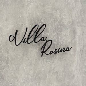 Puede incluir: Un letrero negro con el texto "Villa Rosina" escrito en letra cursiva sobre un fondo gris texturizado.