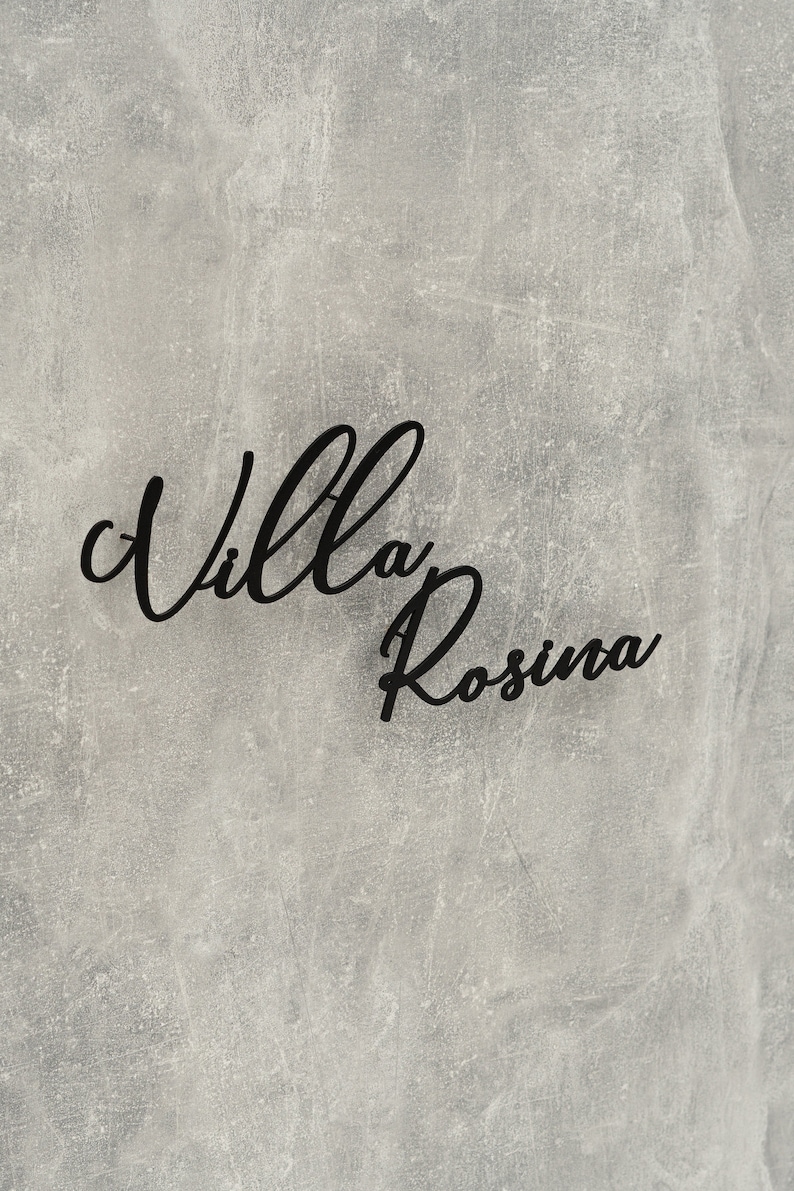 Puede incluir: Un letrero negro con el texto "Villa Rosina" escrito en letra cursiva sobre un fondo gris texturizado.