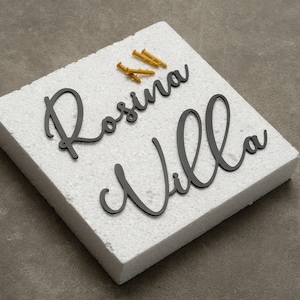 Puede incluir: Tablero de espuma blanco con las palabras "Rosina Villa" escritas en letras cursivas negras. Hay cuatro tornillos dorados en el tablero.