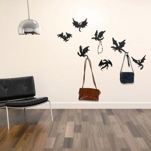 Puede incluir: Un interior moderno presenta ganchos de pared con forma de dragón negro. Dos bolsos marrones y un bolso azul cuelgan de los ganchos. También se muestra un collar con colgante plateado. La pared es de color beige claro y el suelo es de madera.