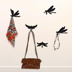 May include: Black metal dragonfly wall hooks. One hook holds a patterned scarf, another a brown purse, and a third a beaded necklace. The hooks are mounted on a white wall, offering a decorative and functional storage solution.