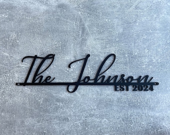Pode incluir: Placa de metal preta com o texto "The Johnson EST 2024" em fonte cursiva. A placa está montada em uma superfície cinza texturizada. Um item decorativo para casa.