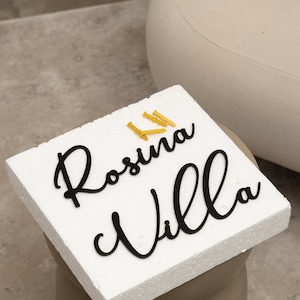 Puede incluir: Tablero de espuma blanco con las palabras "Rosina Villa" escritas en letras cursivas negras. Hay cinco tornillos dorados en el tablero.