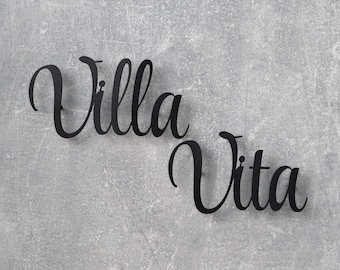 May include: Black metal sign with the words "Villa Vita" in a cursive font. The sign is mounted on a textured, gray background. The letters are connected and have a stylish, elegant appearance.