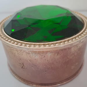 May include: A small, round, silver trinket box with a green, faceted glass top. The box has a beaded rim and a textured surface.