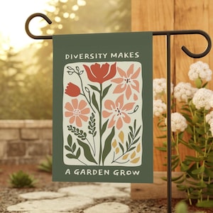 May include: A garden flag with the text "DIVERSITY MAKES A GARDEN GROW" and a floral design in red, pink, and yellow on a dark green background. The flag is displayed on a black metal stand in a garden setting.