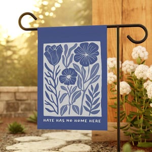 May include: A blue garden flag with a floral design in white and blue. The flag reads "HATE HAS NO HOME HERE" in white text. The flag is hanging on a black metal stand in a garden setting.