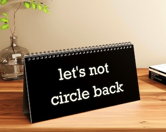 May include: A black desk calendar with the words "let's not circle back" in white. The calendar is on a wooden surface, with a vase of greenery and a stack of books in the background.