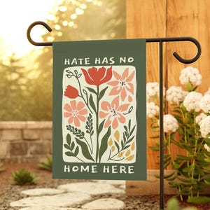 May include: A garden flag with the text "HATE HAS NO HOME HERE" and a floral design in shades of red, pink, and green. The flag is displayed on a black metal stand in a garden setting.