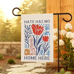May include: A garden flag with the text "HATE HAS NO HOME HERE" and a floral design. The design features red tulips, blue leaves, and a brown flower on a cream background. The flag is displayed on a black metal stand.