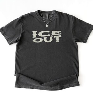 Puede incluir: Una camiseta gris oscuro con el texto "ICE OUT" en una fuente beige claro estilizada. La camiseta tiene mangas cortas y cuello redondo. Se ve un collar plateado con un peque&ntilde;o colgante. La palabra "PEPPER" est&aacute; impresa en la parte inferior.