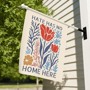 May include: A cream-colored garden flag with the text "HATE HAS NO HOME HERE" and a floral design in red, blue, and orange. The flag is hanging on a white pole attached to a white wall.