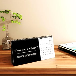 May include: A black and white desk calendar for May with the quote, "There's no 'i' in 'team'. But there are two in 'crisis'" printed on the front. The calendar is on a wooden surface.