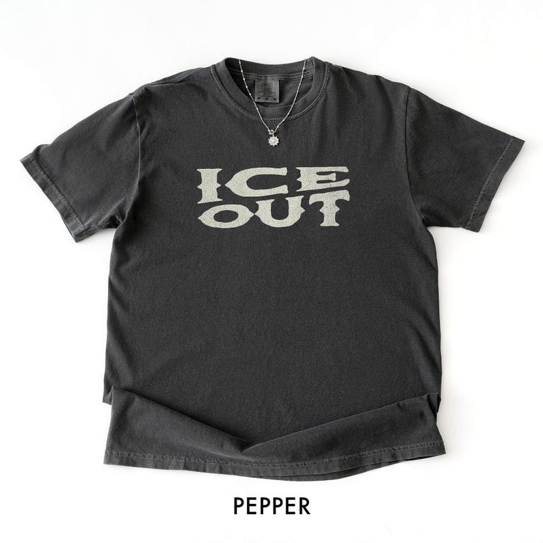 Puede incluir: Una camiseta gris oscuro con el texto "ICE OUT" en una fuente beige claro estilizada. La camiseta tiene mangas cortas y cuello redondo. Se ve un collar plateado con un peque&ntilde;o colgante. La palabra "PEPPER" est&aacute; impresa en la parte inferior.