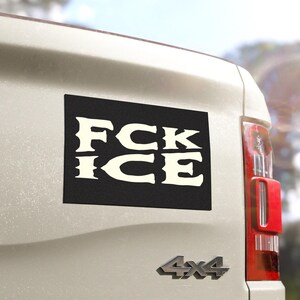 May include: A black rectangular sticker with the words "FCK ICE" in bold, off-white letters is affixed to the back of a white vehicle. The sticker is positioned on the lower left side of the vehicle's rear. The vehicle also has a red tail light.