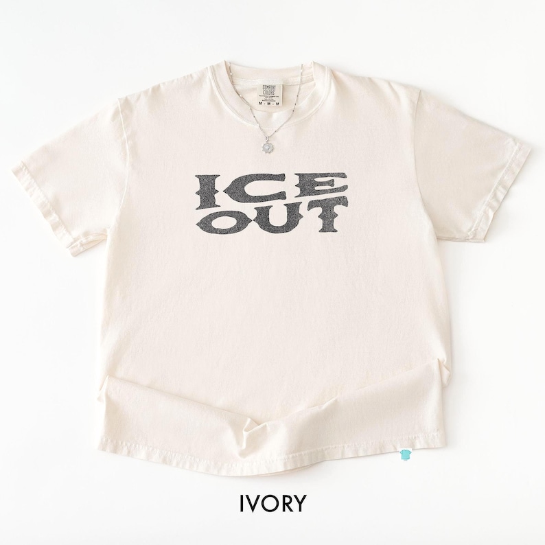 Puede incluir: Camiseta color marfil con el texto "ICE OUT" en gris oscuro, fuente desgastada. La camiseta tiene mangas cortas y cuello redondo. Se ve un collar plateado con un colgante. La palabra "IVORY" est&aacute; impresa en la parte inferior.