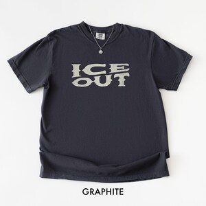 Puede incluir: Camiseta de color grafito con el texto "ICE OUT" en una fuente estilizada. La camiseta tiene mangas cortas y cuello redondo. Se ve un collar plateado con un colgante. La palabra "GRAPHITE" est&aacute; impresa en la parte inferior de la imagen.