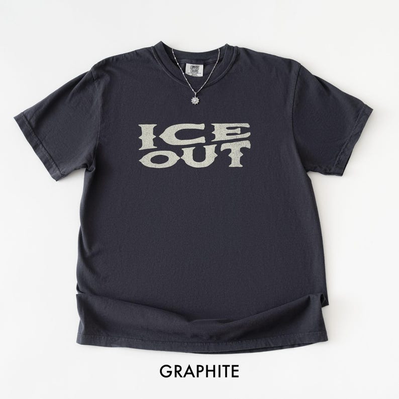 Puede incluir: Camiseta de color grafito con el texto "ICE OUT" en una fuente estilizada. La camiseta tiene mangas cortas y cuello redondo. Se ve un collar plateado con un colgante. La palabra "GRAPHITE" est&aacute; impresa en la parte inferior de la imagen.