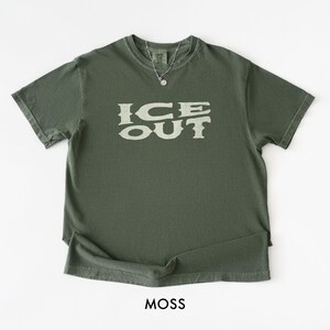 Puede incluir: Una camiseta verde musgo con el texto "ICE OUT" en una fuente desgastada de color blanquecino. La camiseta tiene cuello redondo y mangas cortas. Se ve un collar plateado con un peque&ntilde;o colgante. La palabra "MOSS" est&aacute; impresa en la parte inferior.