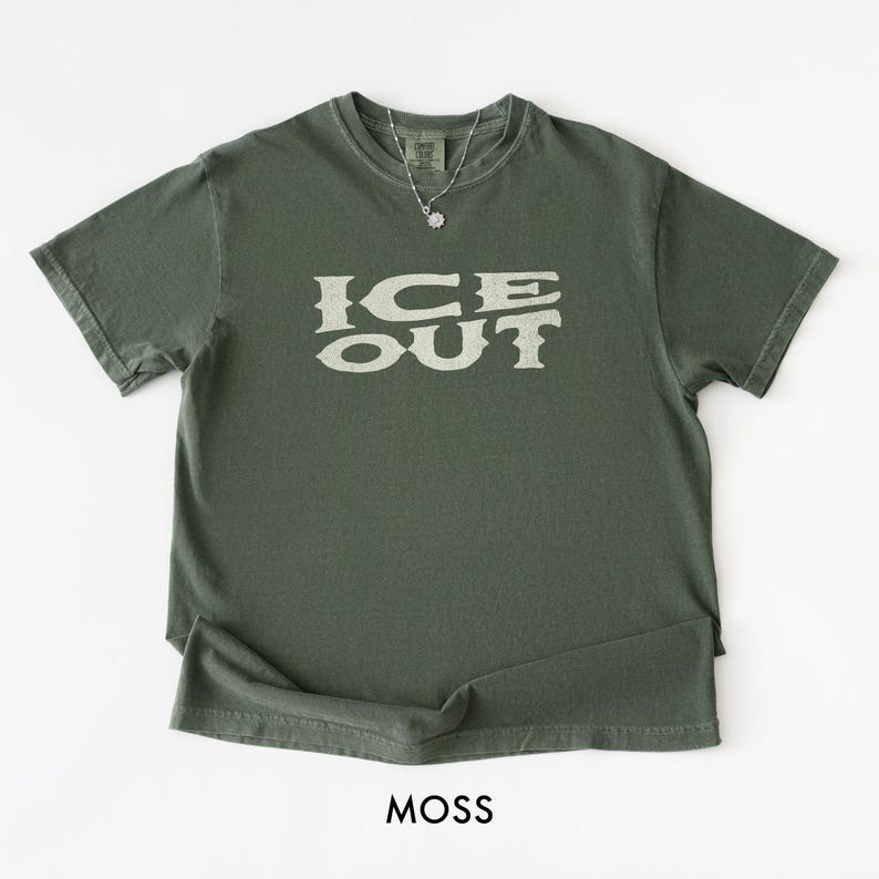 Puede incluir: Una camiseta verde musgo con el texto "ICE OUT" en una fuente desgastada de color blanquecino. La camiseta tiene cuello redondo y mangas cortas. Se ve un collar plateado con un peque&ntilde;o colgante. La palabra "MOSS" est&aacute; impresa en la parte inferior.