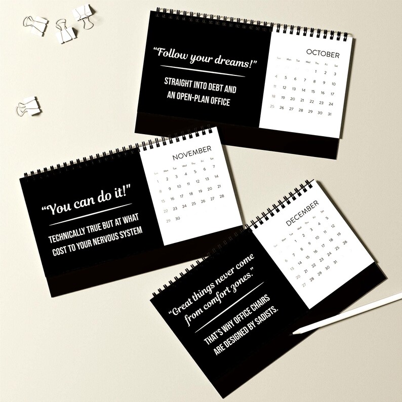 May include: Black and white desk calendars with spiral binding. Each calendar features a month and a humorous quote in white text. The calendars are open to October, November, and December. Paper clips and a pen are also visible.