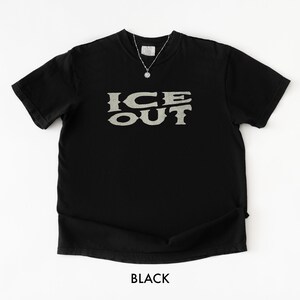 Puede incluir: Camiseta negra con el texto "ICE OUT" en una fuente texturizada de color gris claro. La camiseta tiene cuello redondo y mangas cortas. Se ve un collar plateado con un peque&ntilde;o colgante. La palabra "BLACK" est&aacute; impresa en la parte inferior.