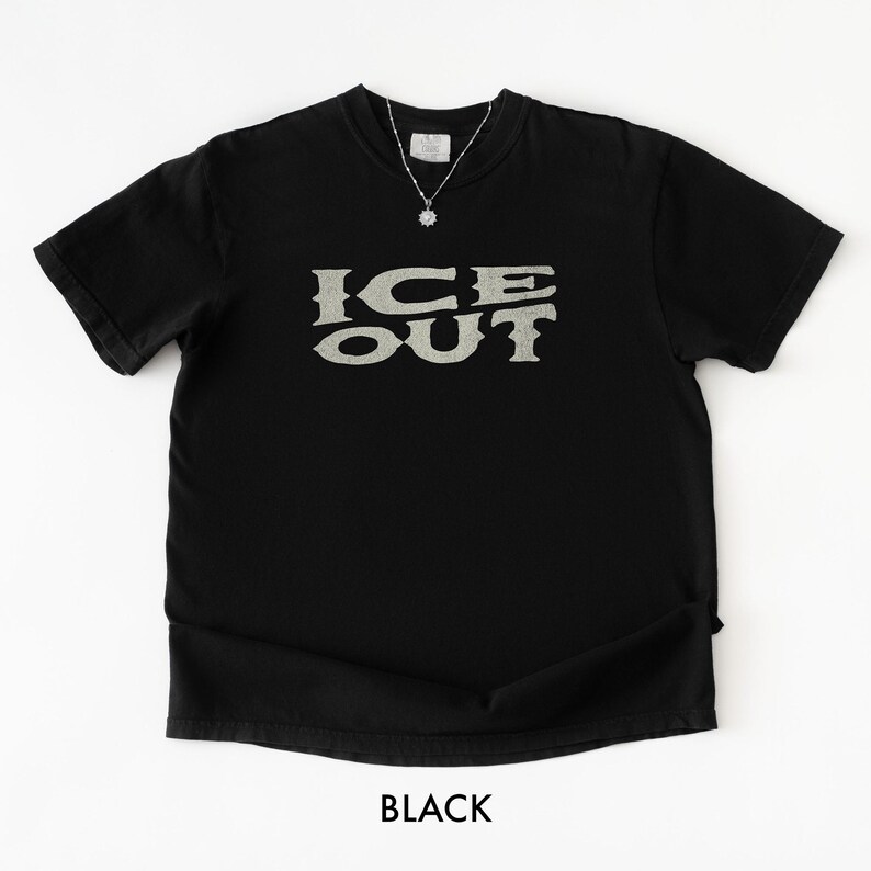 Puede incluir: Camiseta negra con el texto "ICE OUT" en una fuente texturizada de color gris claro. La camiseta tiene cuello redondo y mangas cortas. Se ve un collar plateado con un peque&ntilde;o colgante. La palabra "BLACK" est&aacute; impresa en la parte inferior.