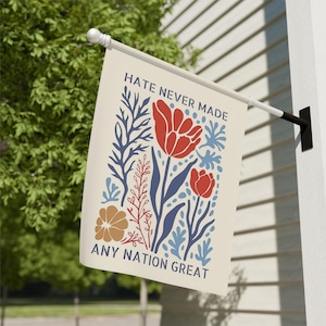 May include: A cream-coloured flag with the text "HATE NEVER MADE ANY NATION GREAT" and a floral design in red, blue, and brown. The flag is mounted on a white pole and is hanging outside.
