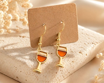 May include: A pair of gold-toned dangle earrings featuring a wine glass charm with a translucent orange-coloured centre. The earrings are attached to a rectangular brown cardboard display card with gold-toned metal ear hooks for pierced ears.