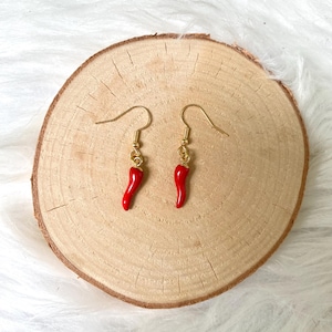 Könnte beinhalten: Ein Paar rote Chili-Pfeffer-Ohrringe mit goldfarbenen Haken. Die Ohrringe werden auf einer runden Holzscheibe mit einem hellen, strukturierten Hintergrund präsentiert. Die Chilischoten haben eine leuchtend rote Farbe.