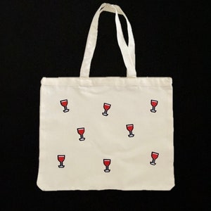 Könnte beinhalten: Eine weiße Canvas-Tragetasche mit einem sich wiederholenden Muster aus roten Weingläsern. Die Tasche hat zwei Henkel.