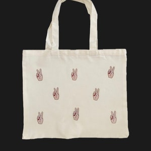 Könnte beinhalten: Eine weiße Canvas-Tragetasche mit einem Peace-Zeichen-Design. Die Peace-Zeichen sind in Pink gestickt.