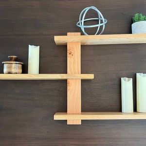 May include: A wooden shelf unit with three shelves. The top shelf has a silver metal orb and a small potted succulent plant. The middle shelf has a small wooden container and a white candle. The bottom shelf has two white candles.